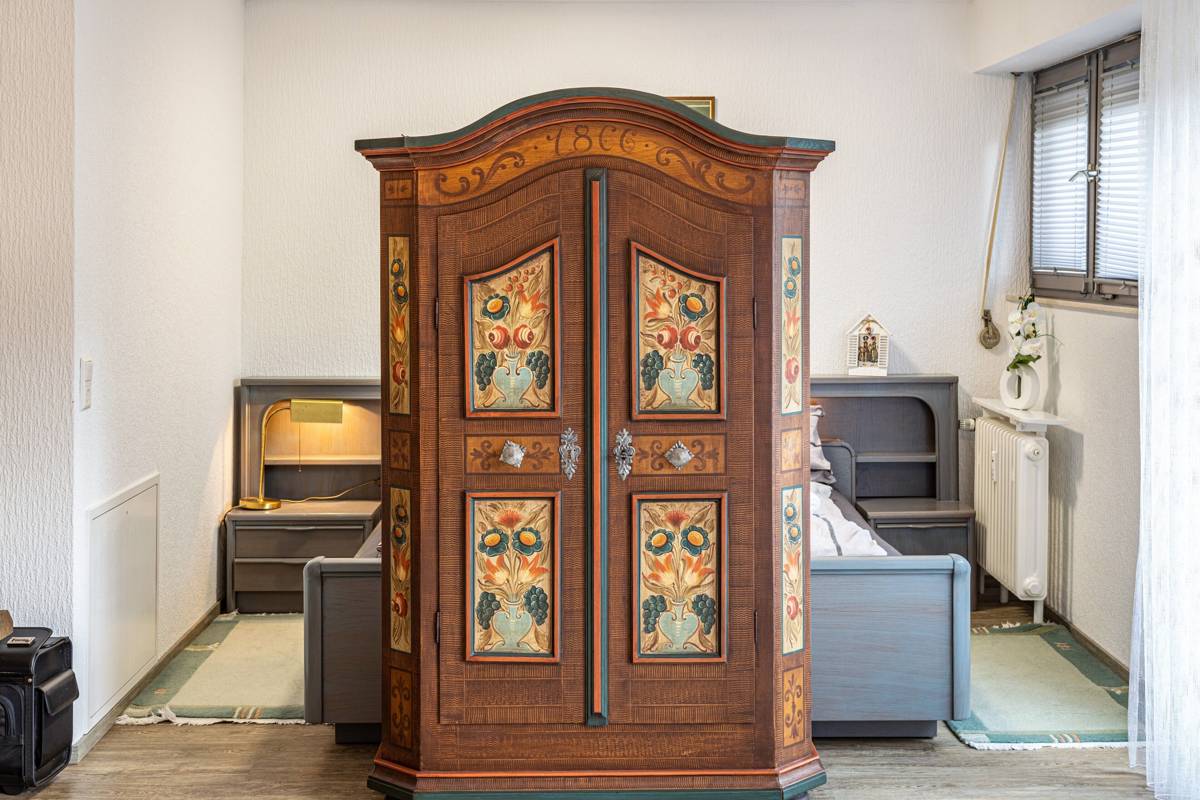A room with a large wooden cabinet with floral designs, a bed, a nightstand, a lamp, and a radiator.