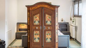 A room with a large wooden cabinet with floral designs, a bed, a nightstand, a lamp, and a radiator.