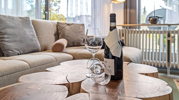 Une table basse en bois avec une bouteille de vin et un verre, un canapé beige et une vue sur l’extérieur.