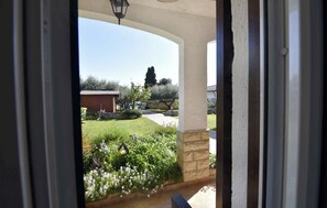 Vista attraverso una porta finestra su un giardino con prato, un piccolo capanno e un albero alto.