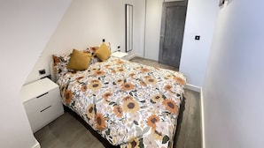Ein Schlafzimmer mit schrägem Dach, einem Bett mit floralem Überwurf, einem Nachttisch, einem Kleiderschrank und einer Tür mit Holzpaneel.