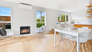 A modern living room with a fireplace, a dining area with a glass table, and a kitchen with white cabinets and a wooden shelf.