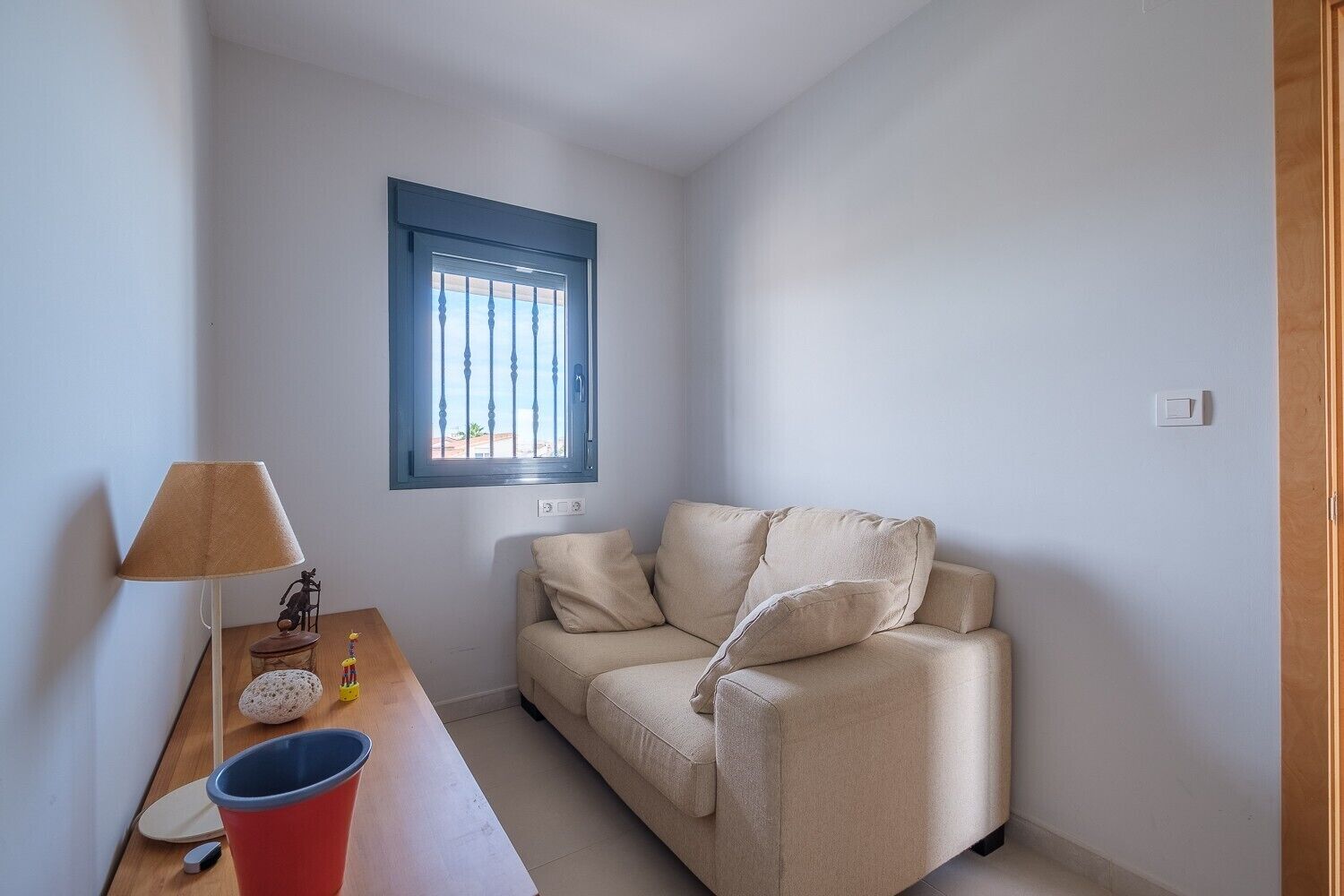 Um quarto pequeno e bem iluminado, com um sofá bege, uma mesa de cabeceira de madeira com um abajur, uma xícara vermelha e uma janela com persianas azuis.