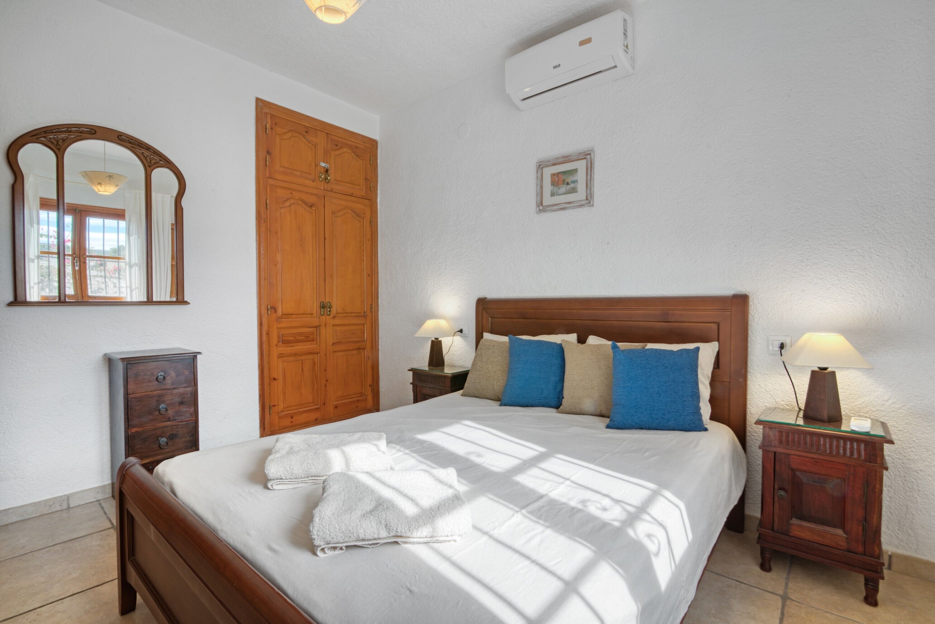 A bedroom with a wooden bed, two bedside tables with lamps, a wardrobe, a mirror, and an air conditioning unit.