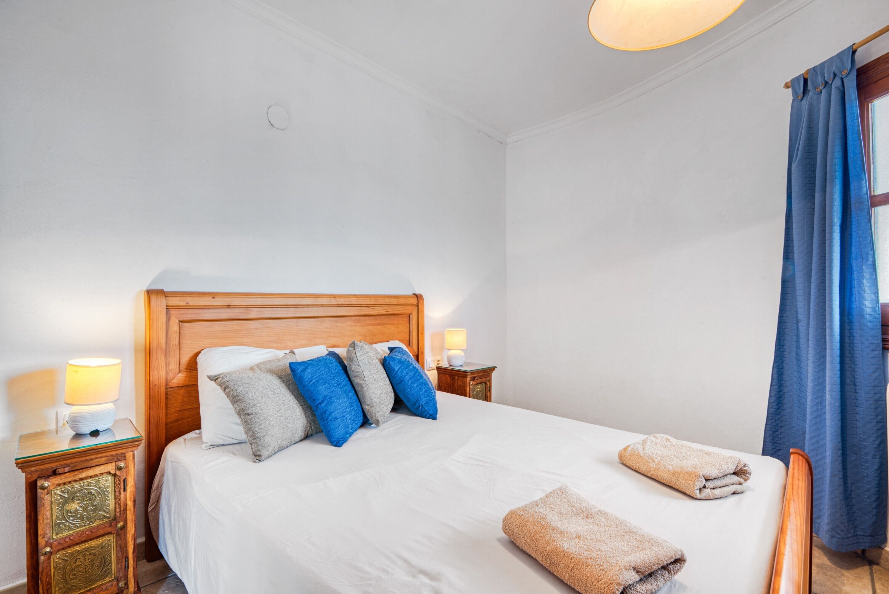 A neatly made bed with pillows, a wooden headboard, bedside lamps, and a window with blue curtains.