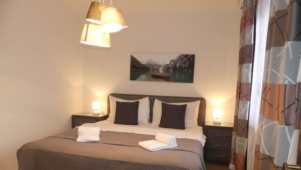 A neatly made bed with a brown bedspread, two bedside lamps, a painting on the wall, and a patterned curtain. - Apartment Siegl by Interhome (Zell am See)