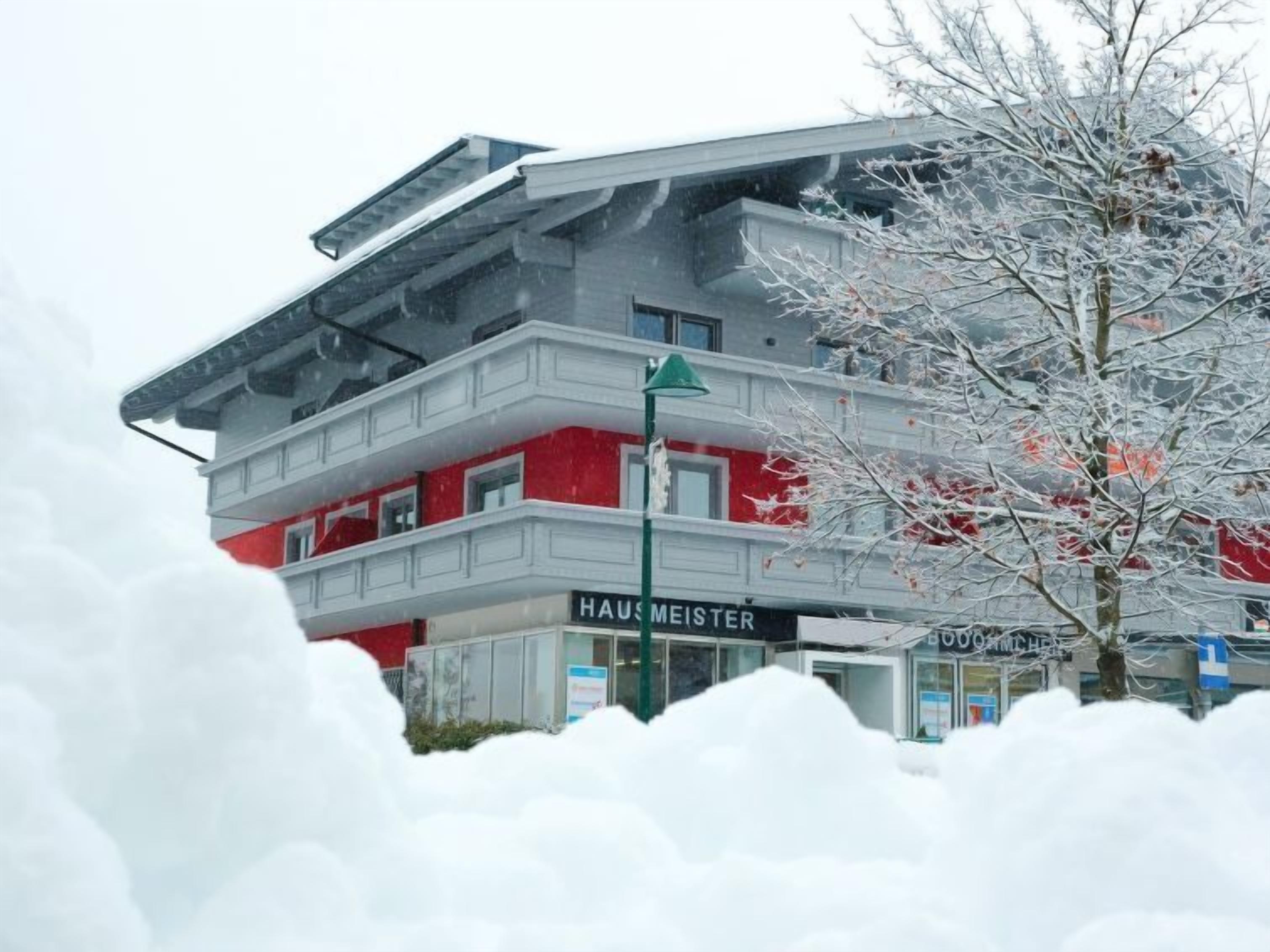 Ein mehrstöckiges Gebäude mit roter und weißer Fassade, einem sichtbaren Schild mit der Aufschrift „HAUS MEISTER“ und einem mit Schnee bedeckten Baum im Vordergrund.