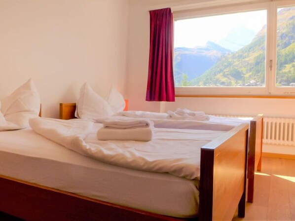 A neatly made twin bed with white linens and pillows, a wooden headboard, and a view of a mountainous landscape through a window with a red curtain.