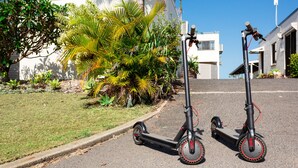 Two electric scooters parked on a street with a building and palm trees in the background. - One bedroom beachfront apartment (Yeppoon)