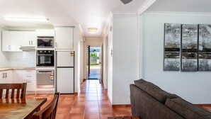A modern kitchen with white cabinets, a dining table with chairs, and a gray sofa in a bright living area. - One bedroom beachfront apartment (Yeppoon)