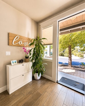 Une pièce avec un plancher en bois, une commode blanche, une grande plante en pot, un panneau fixé au mur et une vue sur un arbre et la rue, à l’extérieur.