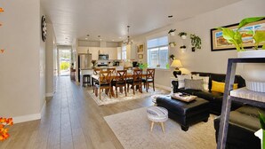 Une cuisine moderne avec des meubles blancs, un coin repas avec des chaises en bois, un canapé en cuir noir et une petite table d’appoint avec une lampe.