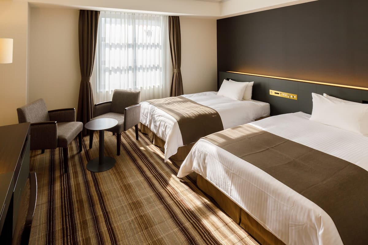 Hotellihuone, jossa on kaksi sänkyä, työpöytä, tuoli ja ikkuna, jossa on verhot.