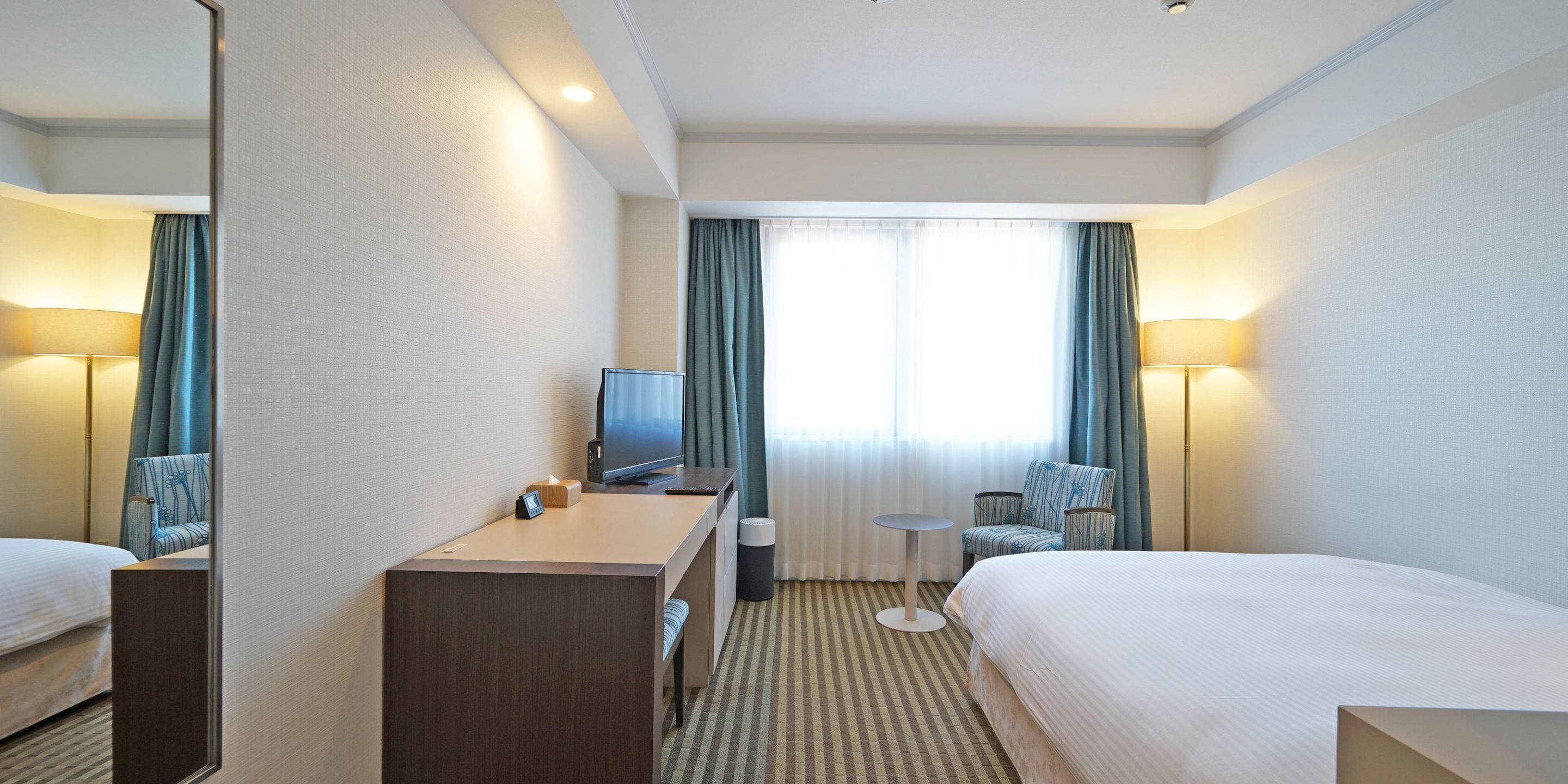 A hotel room with a bed, a desk with a computer, a chair, a TV, and a window with curtains.