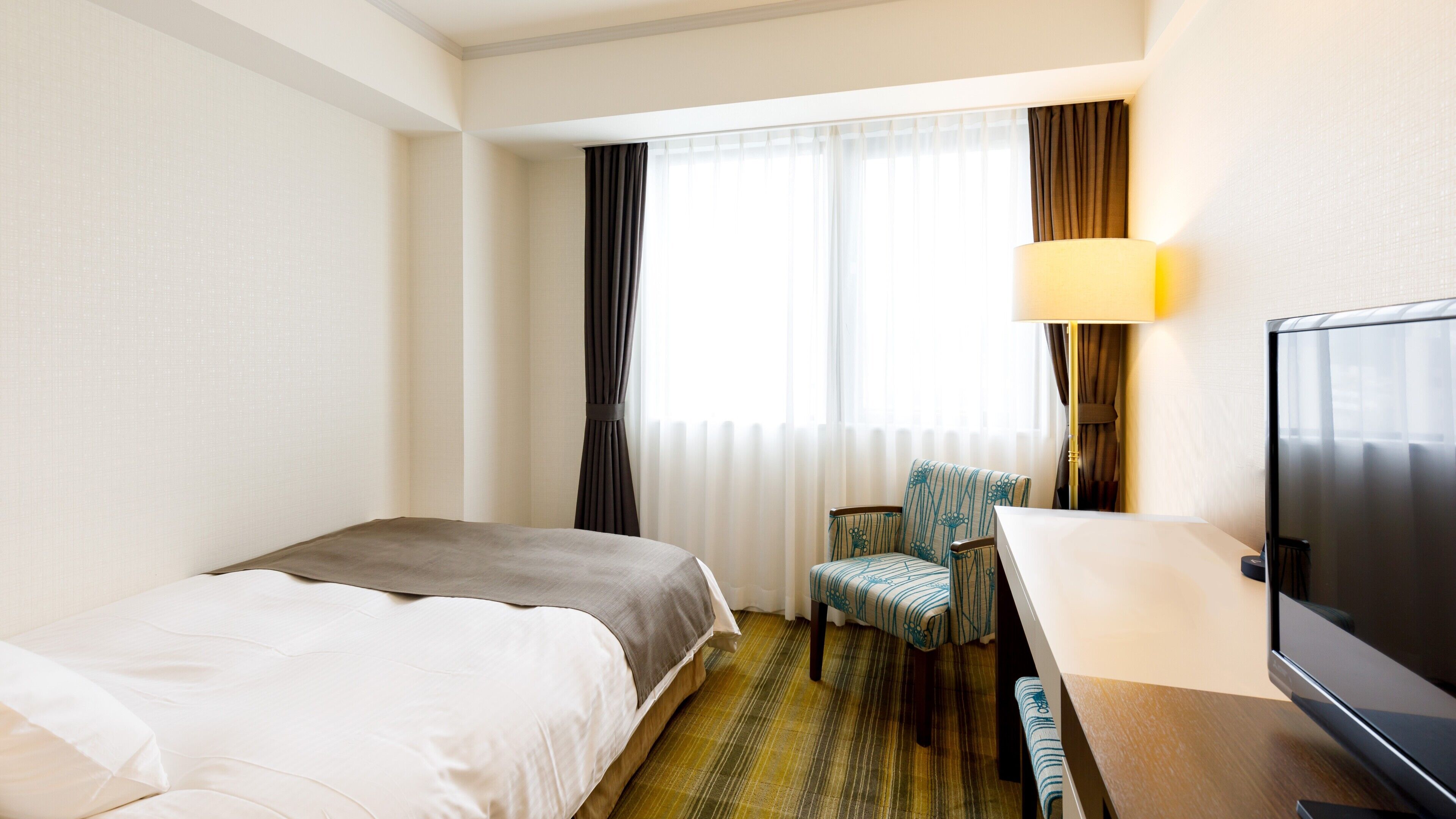 A hotel room with a bed, a desk with a chair, a television, and a window with curtains.