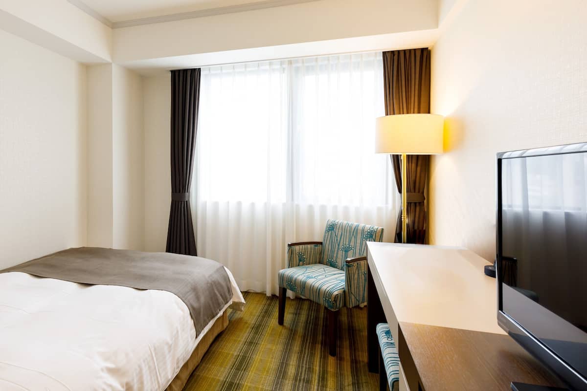Hotellihuone, jossa on sänky, työpöytä tuolilla, televisio ja ikkuna, jossa on verhot.