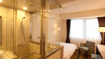 Habitación de hotel con ducha de vidrio, cama, silla y escritorio.
