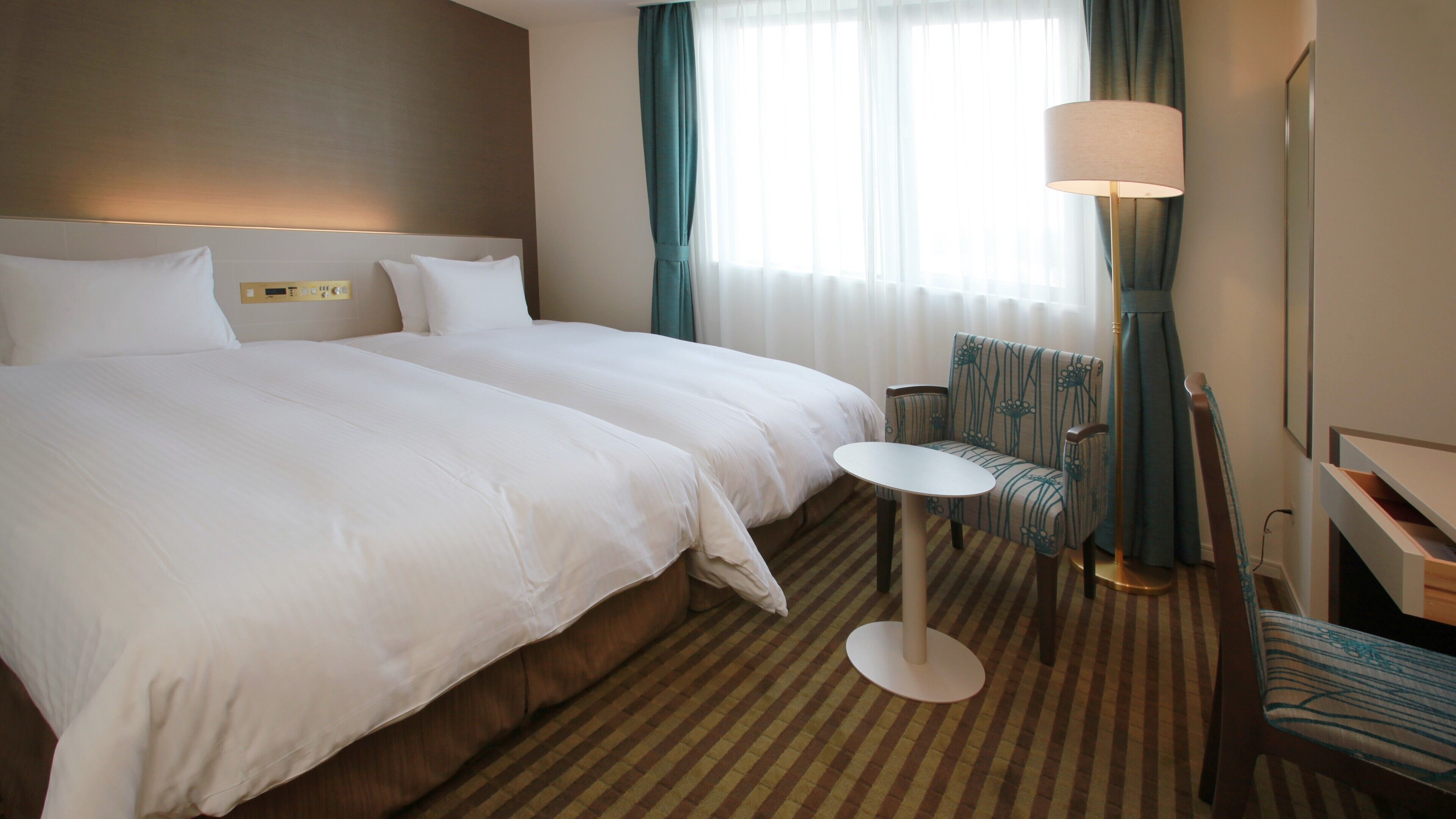A hotel room with two beds, a small round table, a chair, a lamp, and a window with curtains.