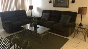 Una sala de estar con dos sofás marrón oscuro, una mesa de centro de vidrio, una planta en maceta y dos lámparas de pared.