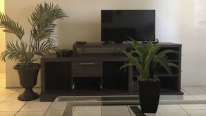 Una sala de estar moderna con un televisor de pantalla plana sobre un mueble negro, dos plantas en macetas y piso de baldosas.