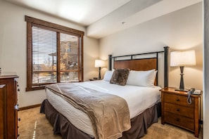 A bedroom with a large bed, two bedside lamps, a wooden nightstand, and a window with blinds.