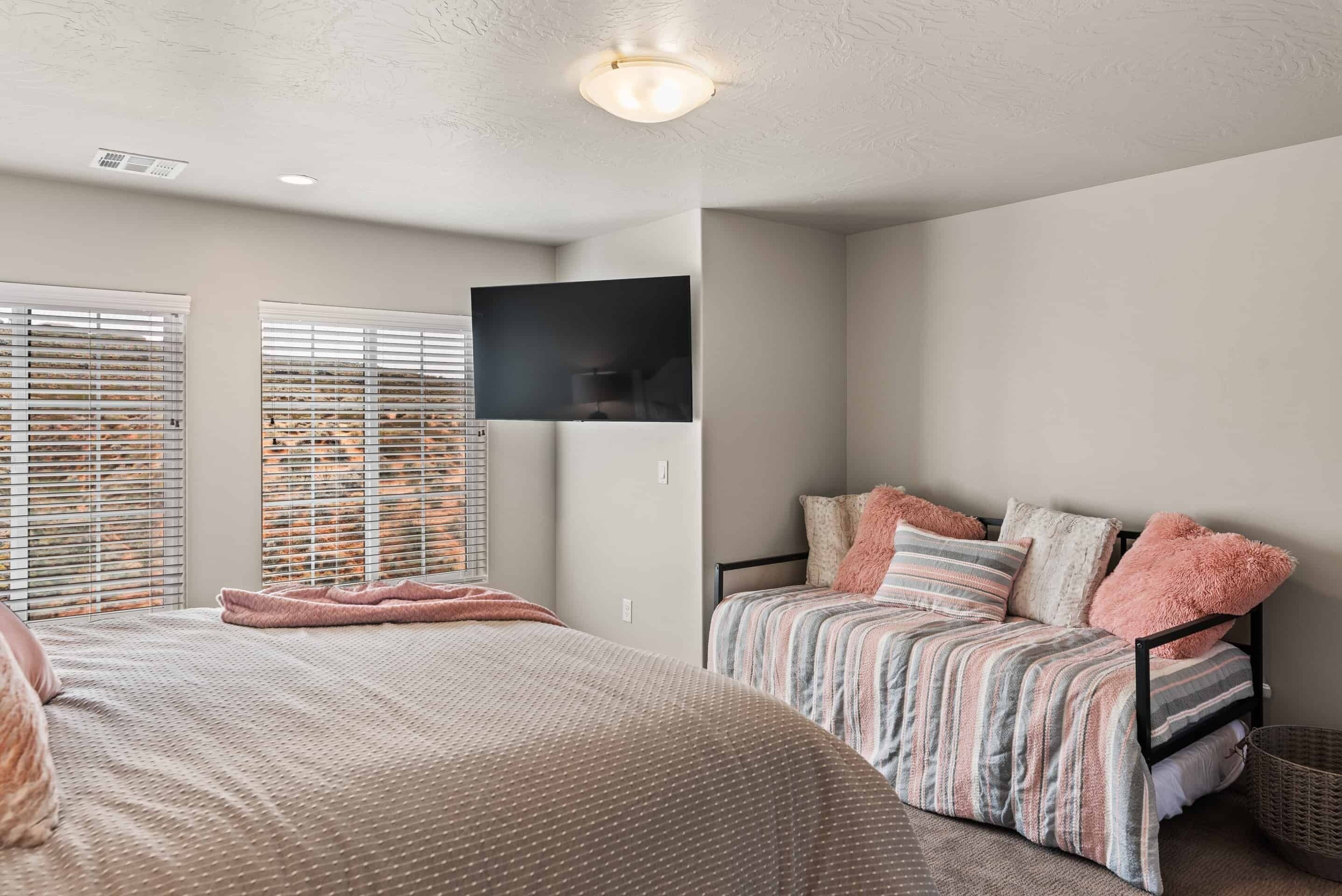 A bedroom with a bed, a sofa, a TV mounted on the wall, and a window with blinds.