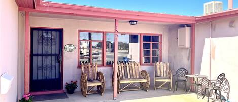 A building with a pink facade, a black door, and a window with a view of the street. There are wooden chairs and a metal table outside.