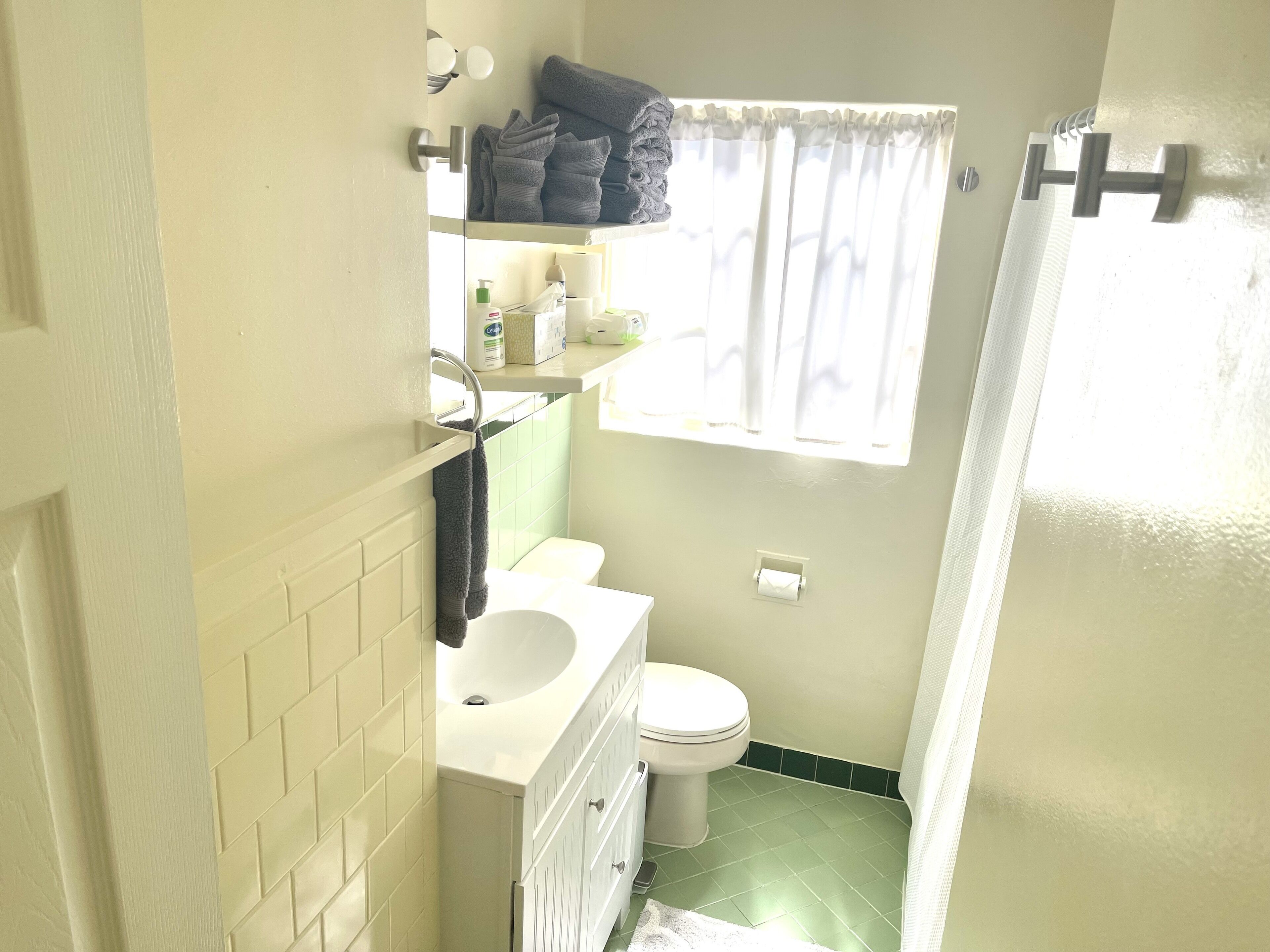 Et lille badeværelse med en hvid vask, toilet og grønne fliser. Der er et vindue med en hvid gardin samt en hylde med håndklæder og toiletartikler.