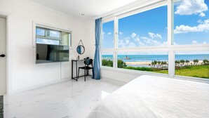 A modern bedroom with a large window offering a beach view, a flat-screen TV, a small desk, and a chair. - Hi Yeongjin Spa Pension (Gangneung)