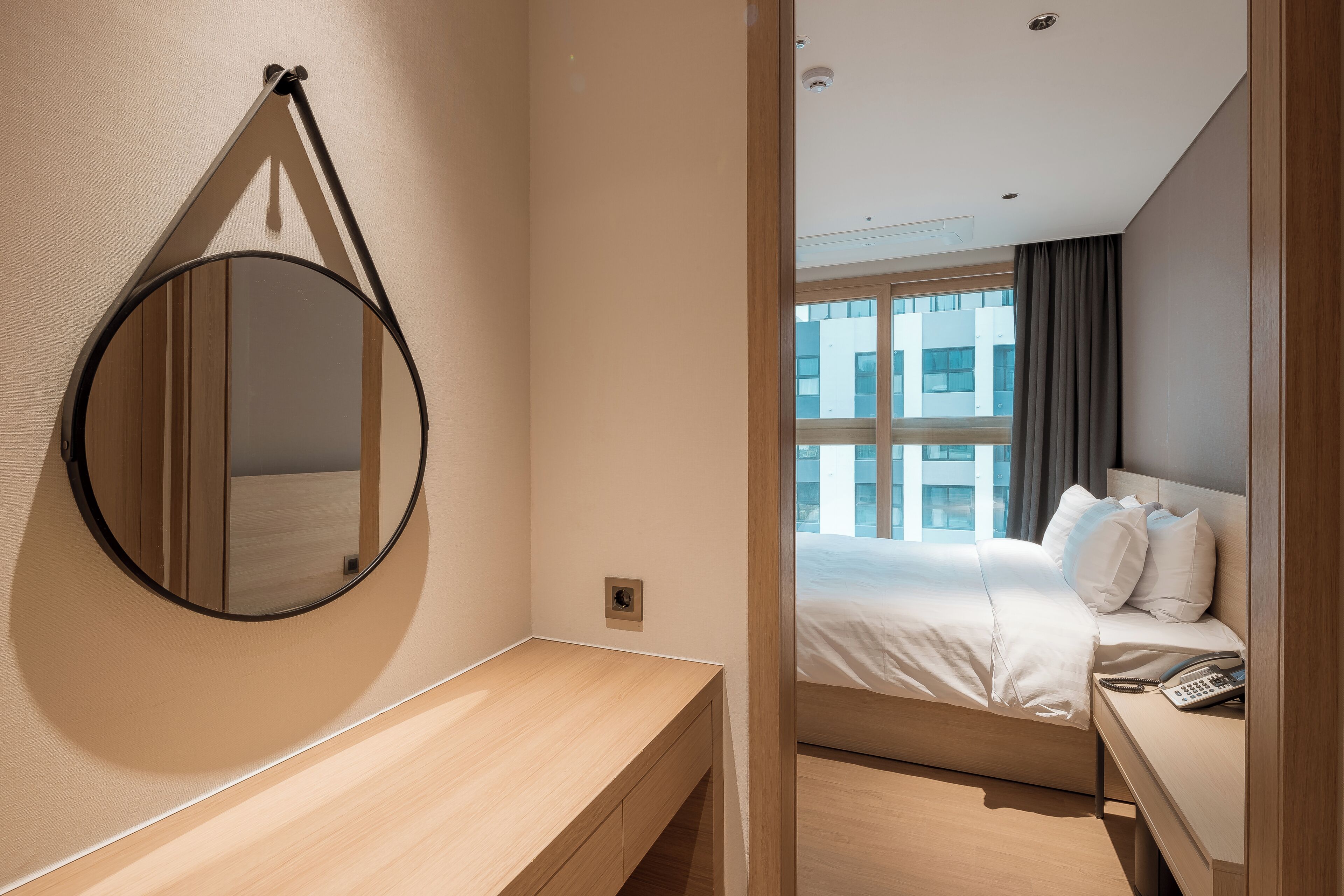 Habitación de hotel con mesita de noche de madera, un espejo redondo, cama con ropa blanca y vistas a edificios a través de una ventana.
