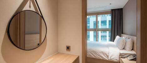 Habitación de hotel con mesita de noche de madera, un espejo redondo, cama con ropa blanca y vistas a edificios a través de una ventana.