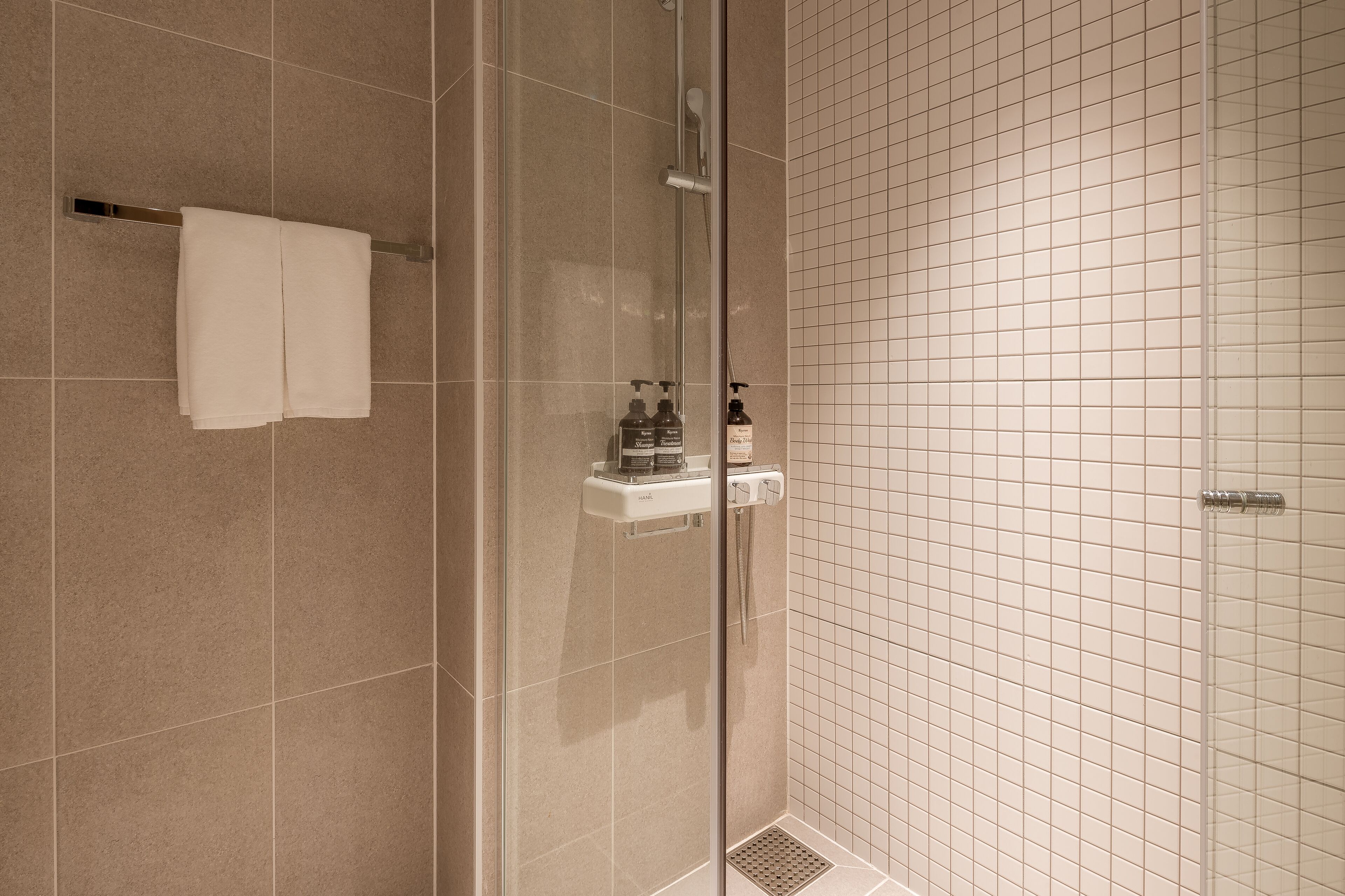 Un baño con cabina de ducha de vidrio, un perchero con dos toallas, un dispensador de jabón y un pequeño estante con artículos de aseo.