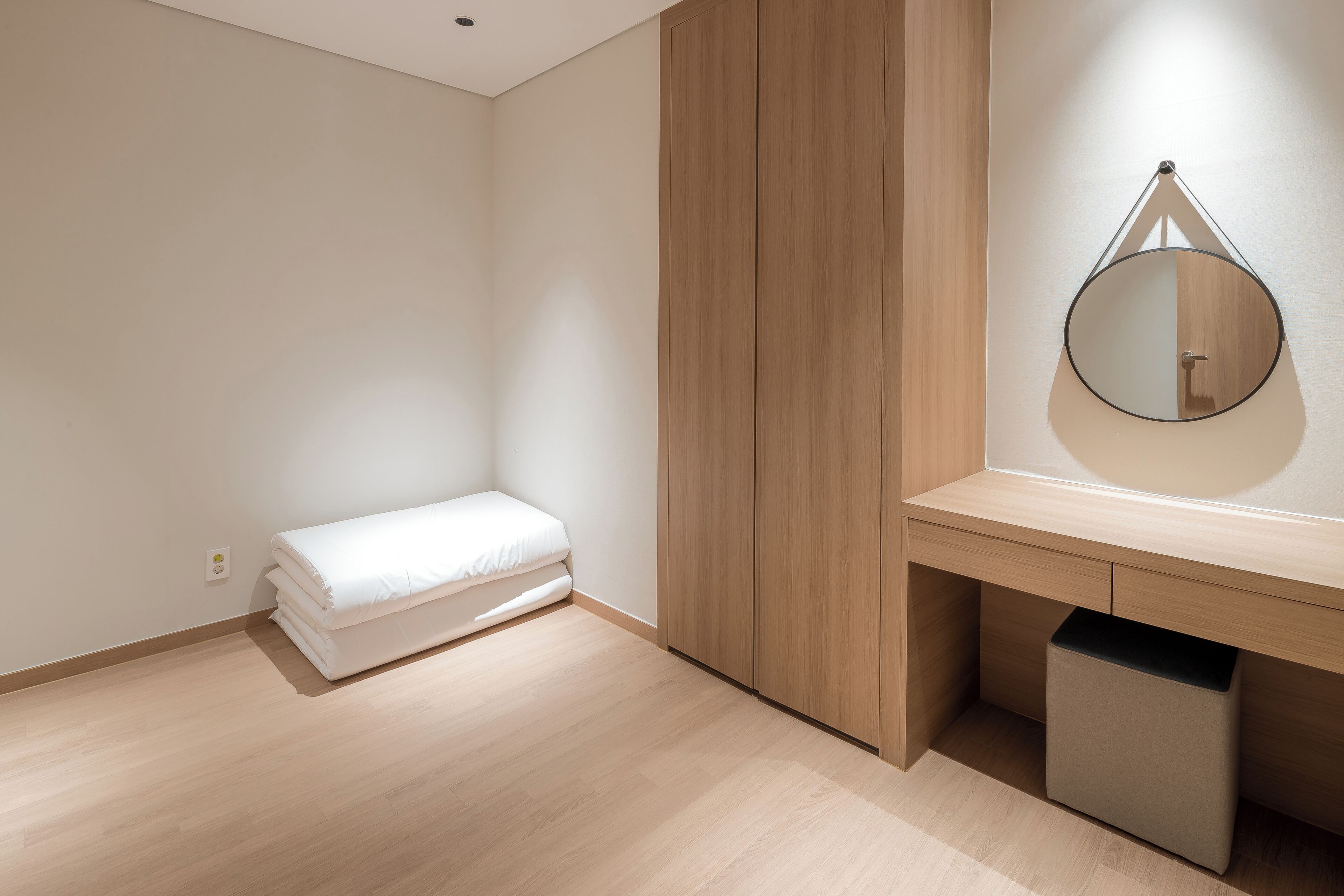 Ein schlicht eingerichteter Raum mit einem Bett, einem Holzschrank, einem runden Spiegel und einer kleinen Mülltonne.