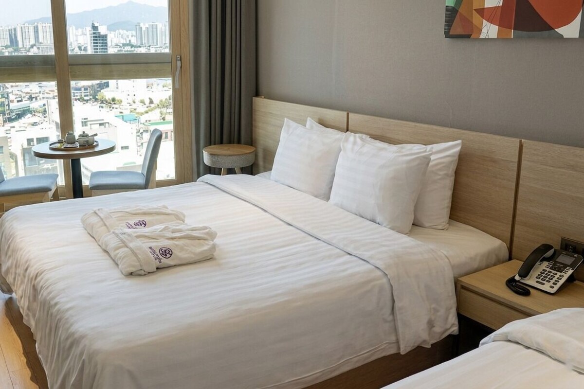 Habitación de hotel con una cama, una mesita de noche con un teléfono, una pequeña mesa redonda con una silla y vistas a la ciudad a través de grandes ventanales.
