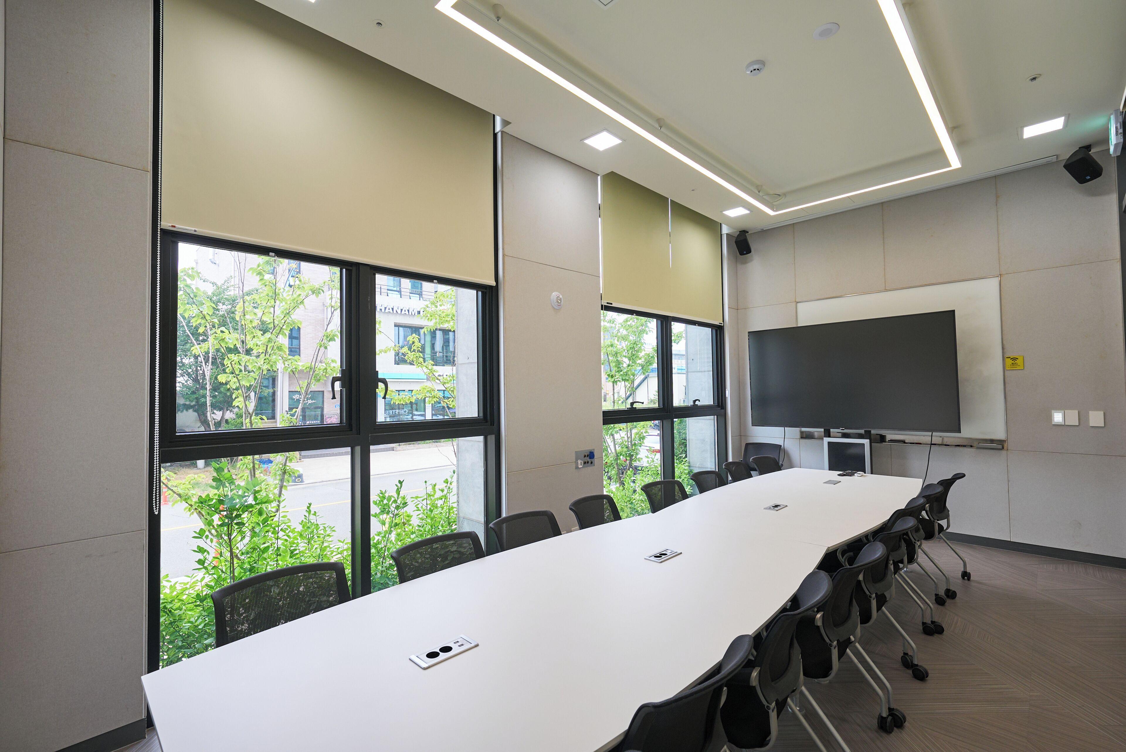 A conference room with a long table, chairs, a projector screen, and large windows with a view of buildings and trees.