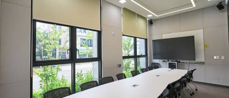 A conference room with a long table, chairs, a projector screen, and large windows with a view of buildings and trees.