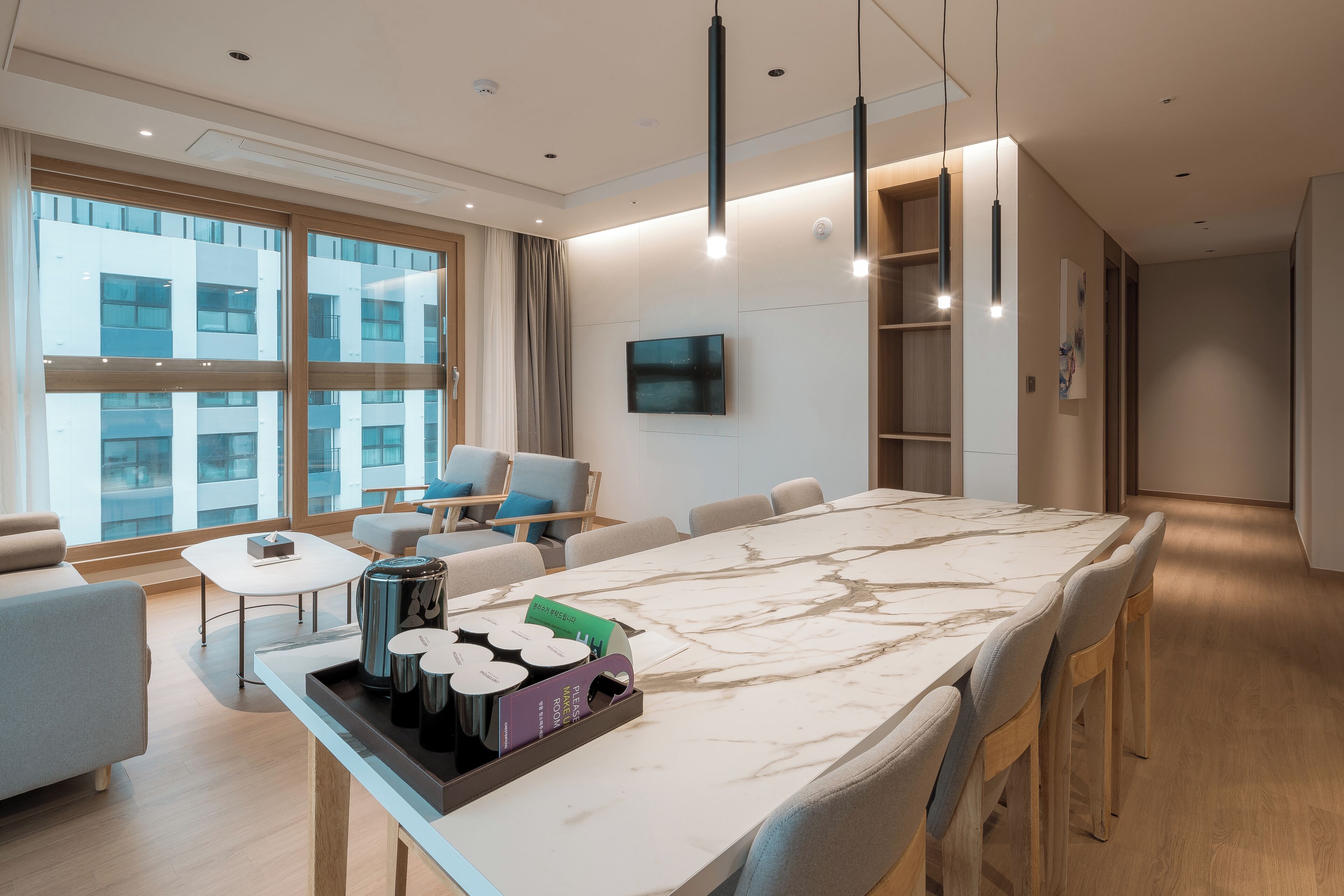 Una habitación de hotel moderna con una amplia mesa de comedor de mármol, asientos cómodos, un televisor de pantalla plana y grandes ventanales con vistas a la ciudad.