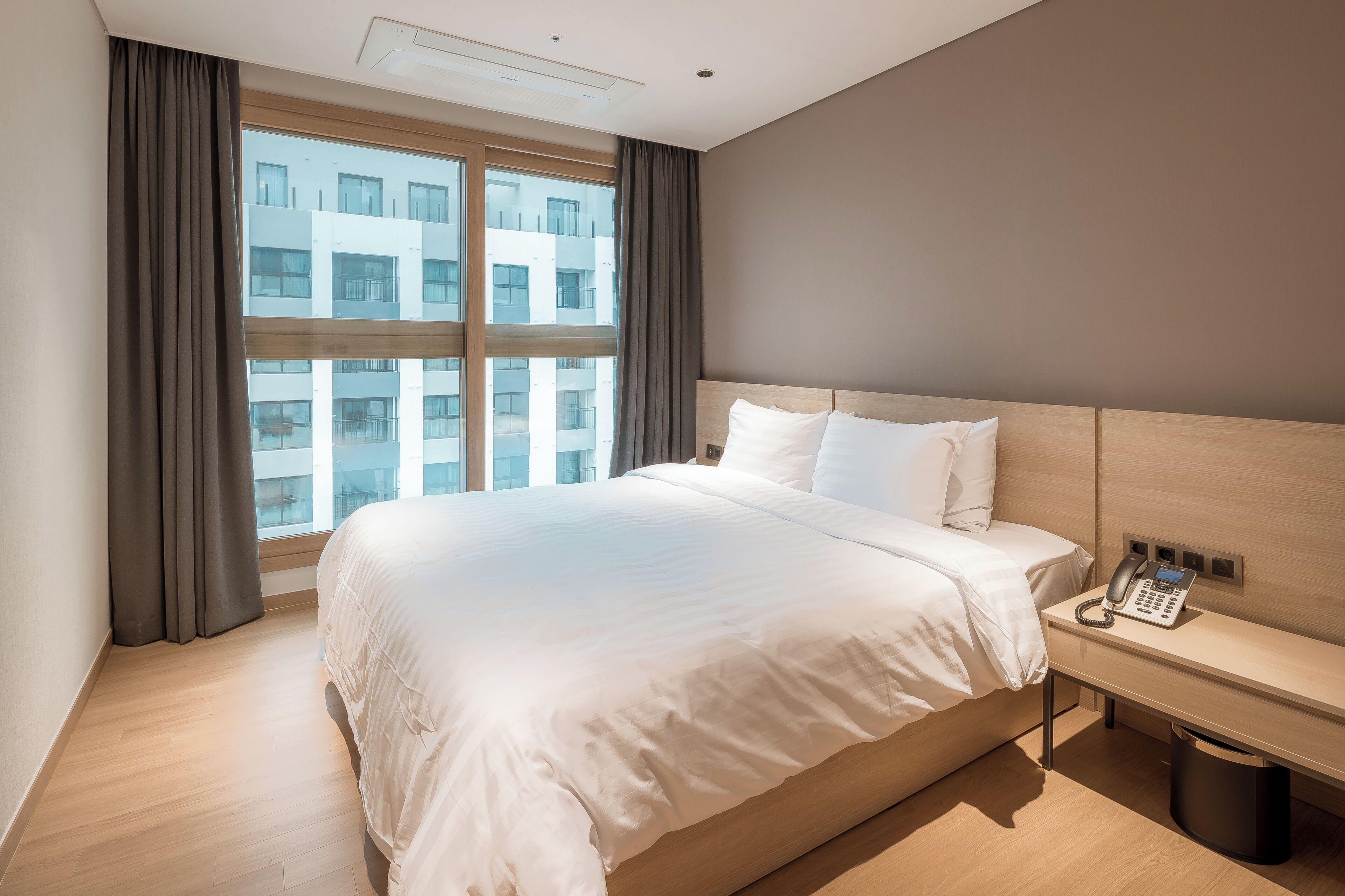 A hotel room with a large bed, a bedside telephone, and a view of a building through the window.