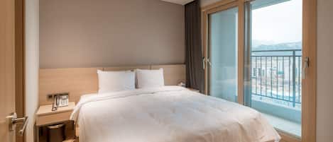 A modern hotel room with a large bed, a bedside telephone, a small trash bin, and a view of the city through a large window.