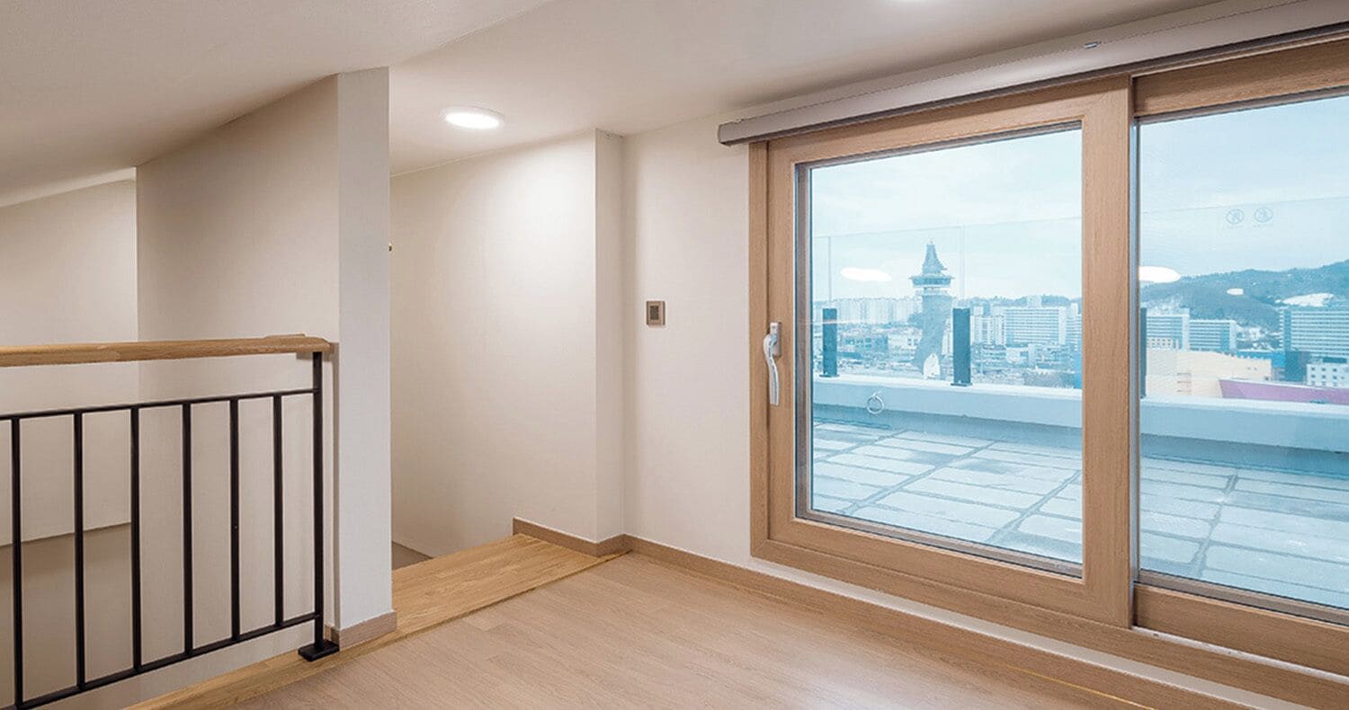 A room with a view of a cityscape through a large window, a wooden floor, and a black metal railing.