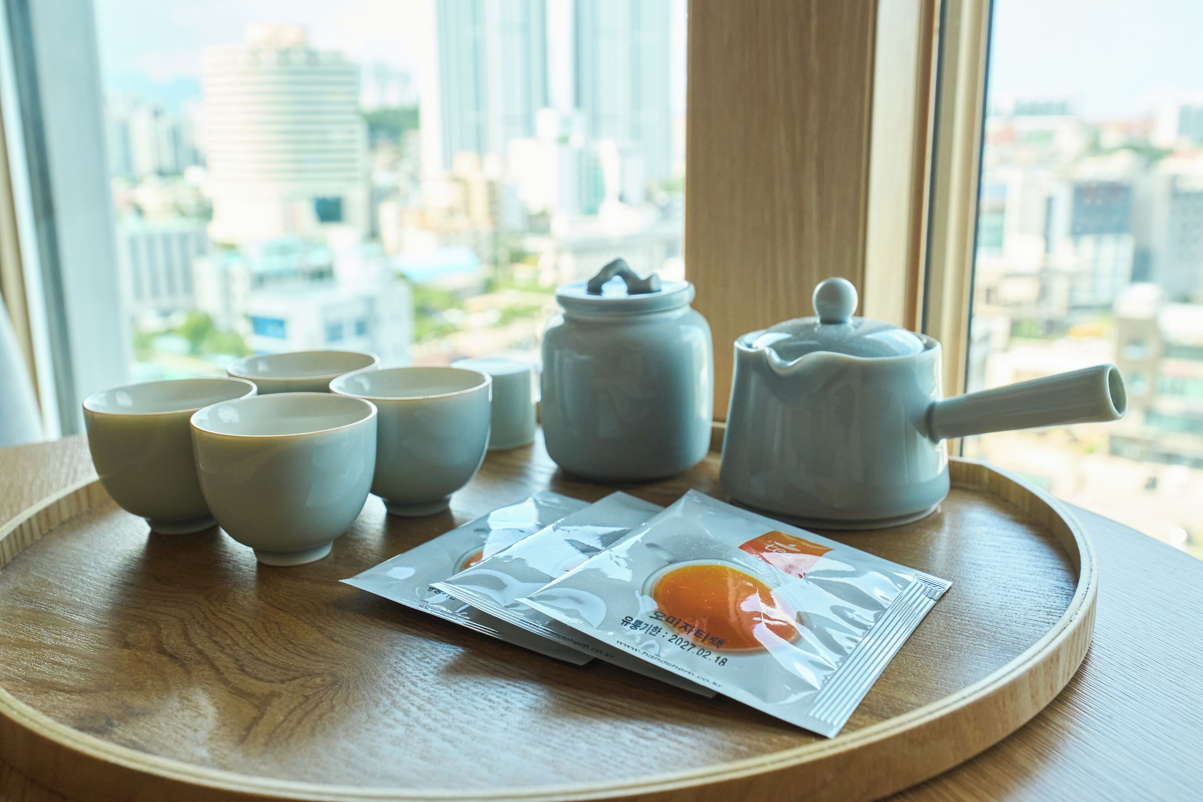 A tray with tea cups, a teapot, and a tea packet on a wooden surface near a window with a city view.