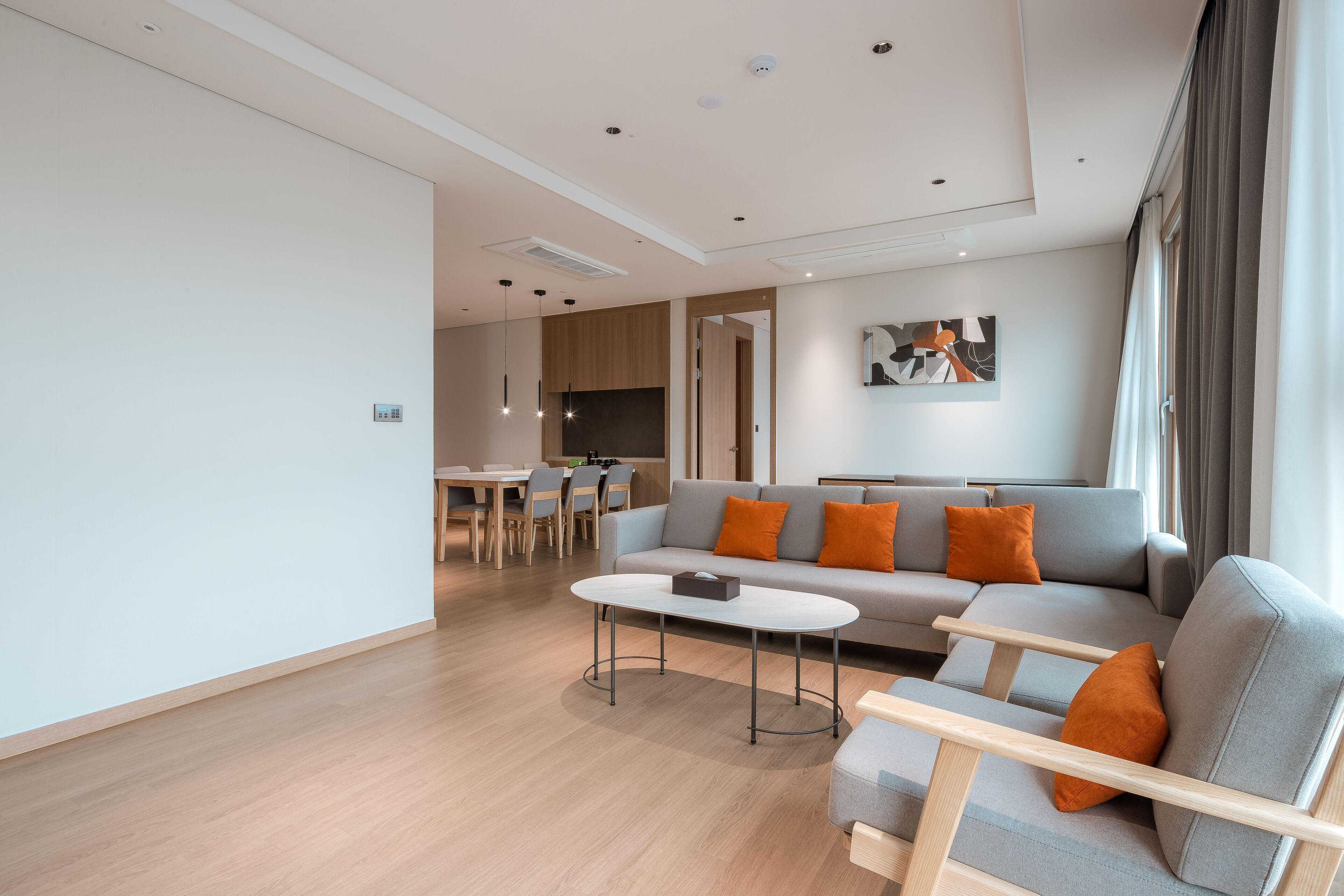 Un salon moderne avec un canapé gris, des coussins orange, une table basse ronde et un coin repas à l’arrière-plan.