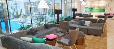 En moderne hotellobby med et stort siddeområde, glasvægge og udsigt til en pool.