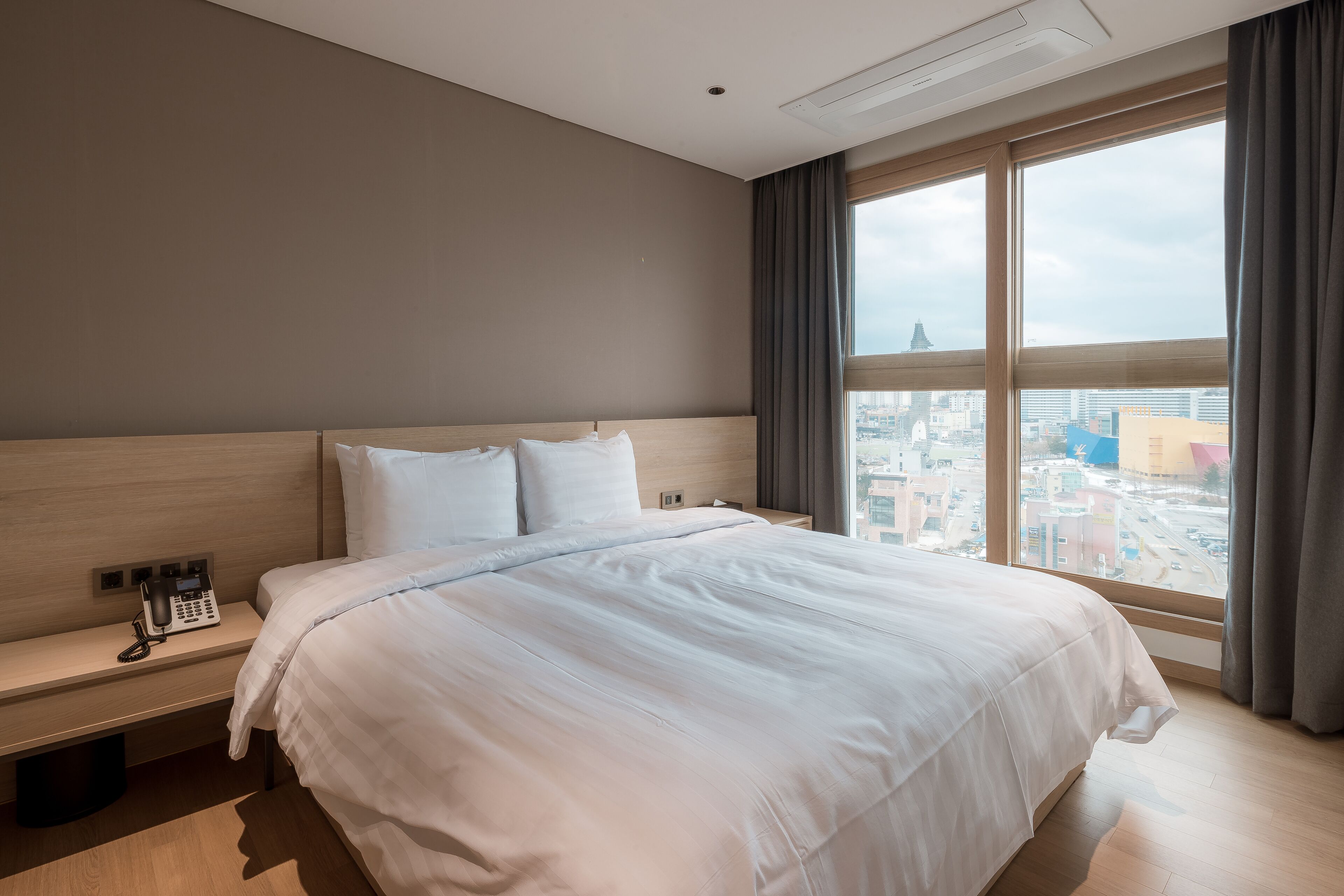 A hotel room with a large bed, a bedside telephone, and a city view through the window.