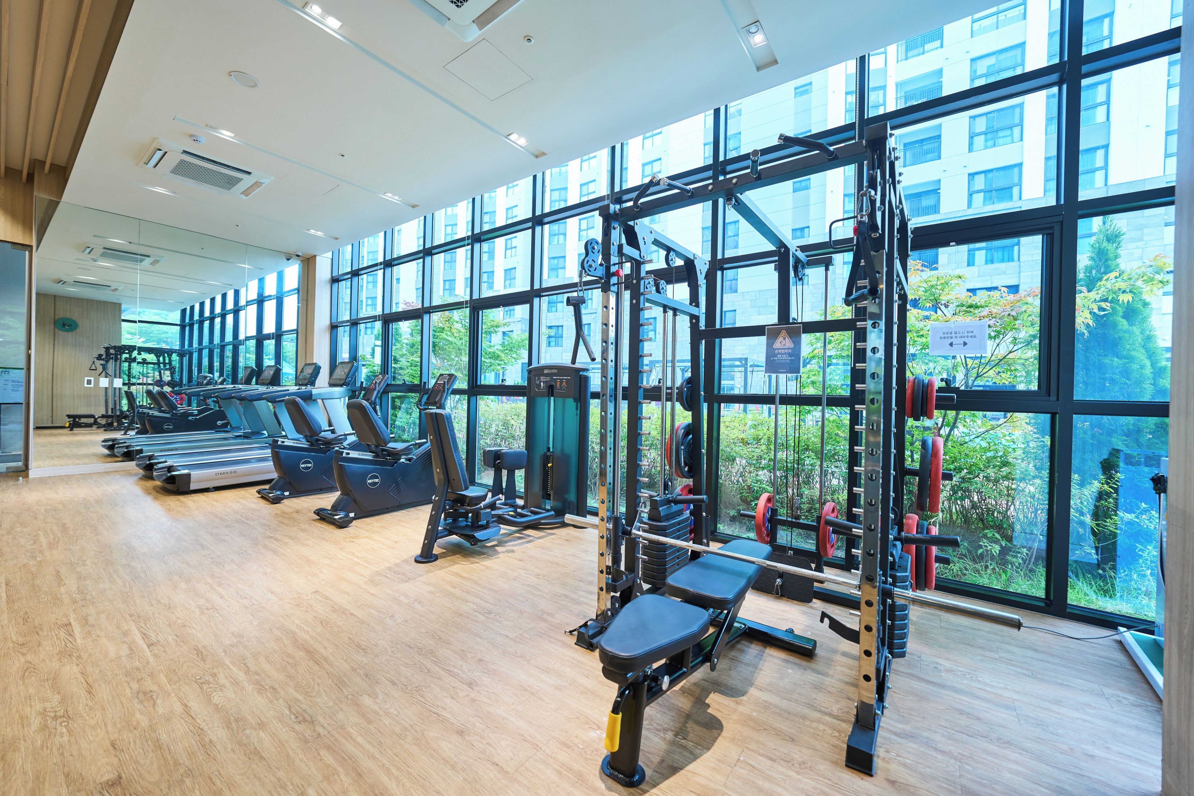 Un gimnasio con cintas de correr, máquinas de pesas y un amplio ventanal con vistas a edificios y árboles.