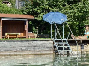 Un muelle de madera con una sombrilla azul, una escalera y una persona descansando junto al agua.