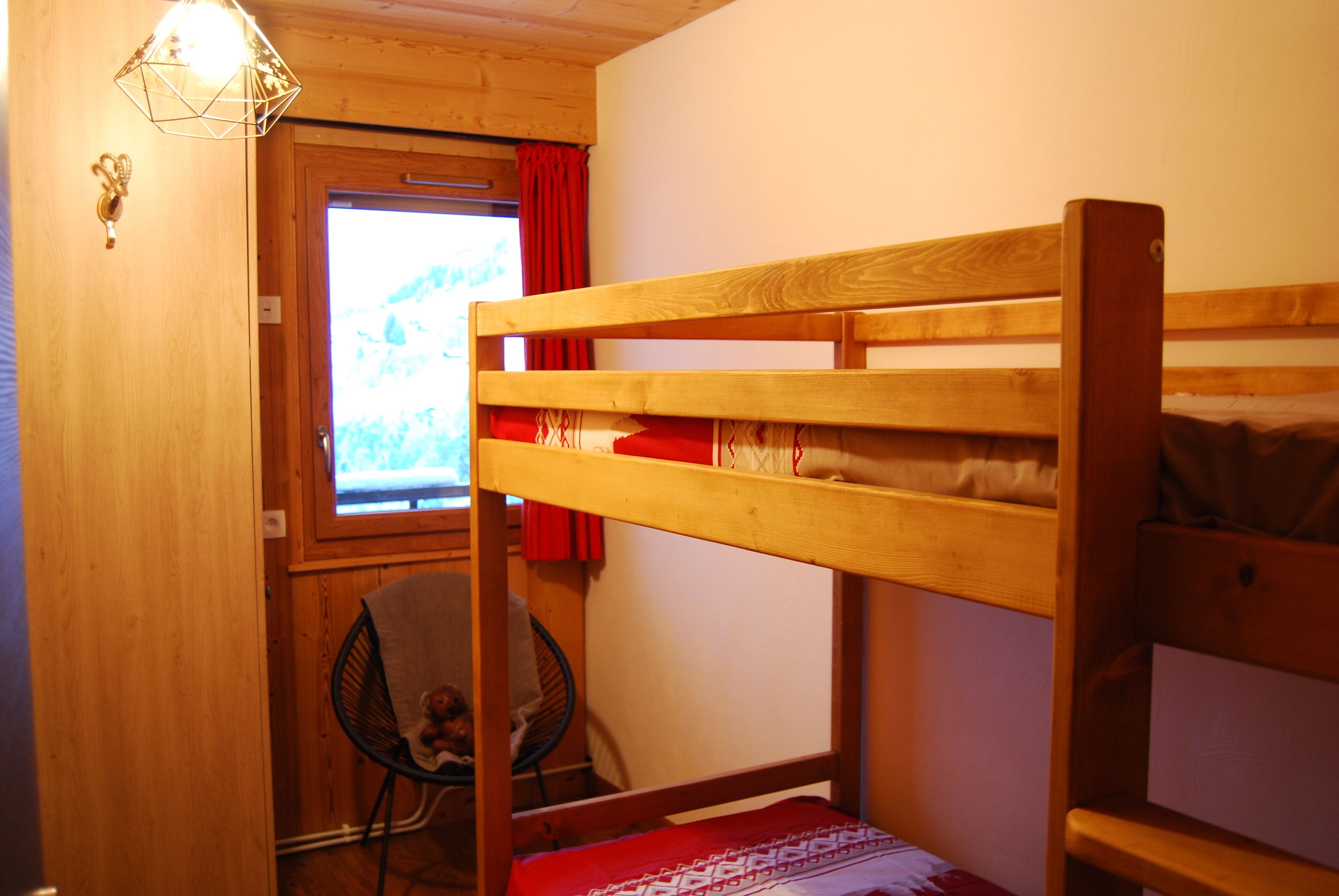 A wooden bunk bed with a window and red curtains, a chair, and a small table.