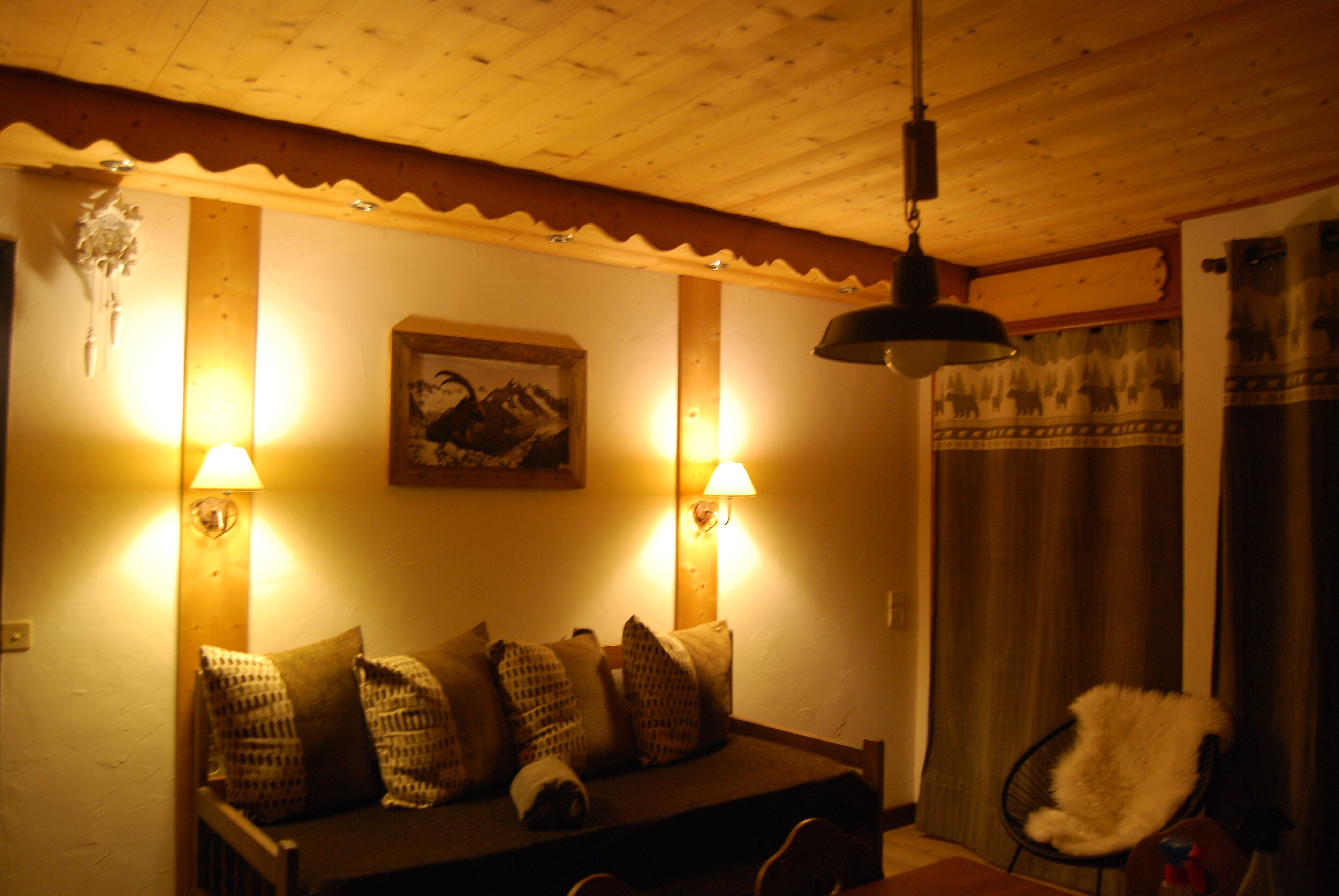 A cozy room with a wooden ceiling, a sofa with patterned cushions, two wall-mounted lamps, and a framed picture on the wall.
