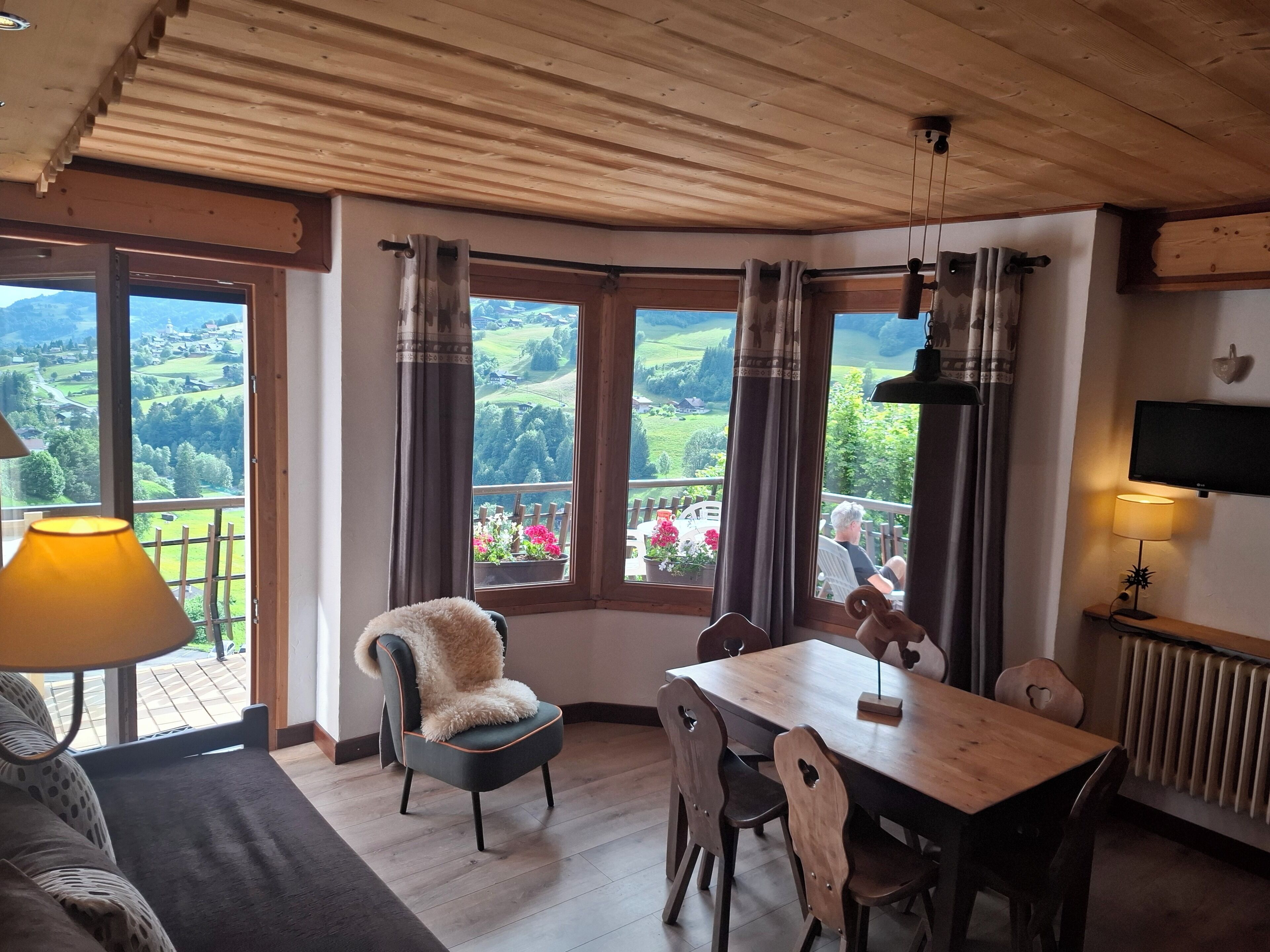 Een gezellige kamer met houten plafond, een eettafel met stoelen, een zachte fauteuil, een lamp en een televisie aan de muur.