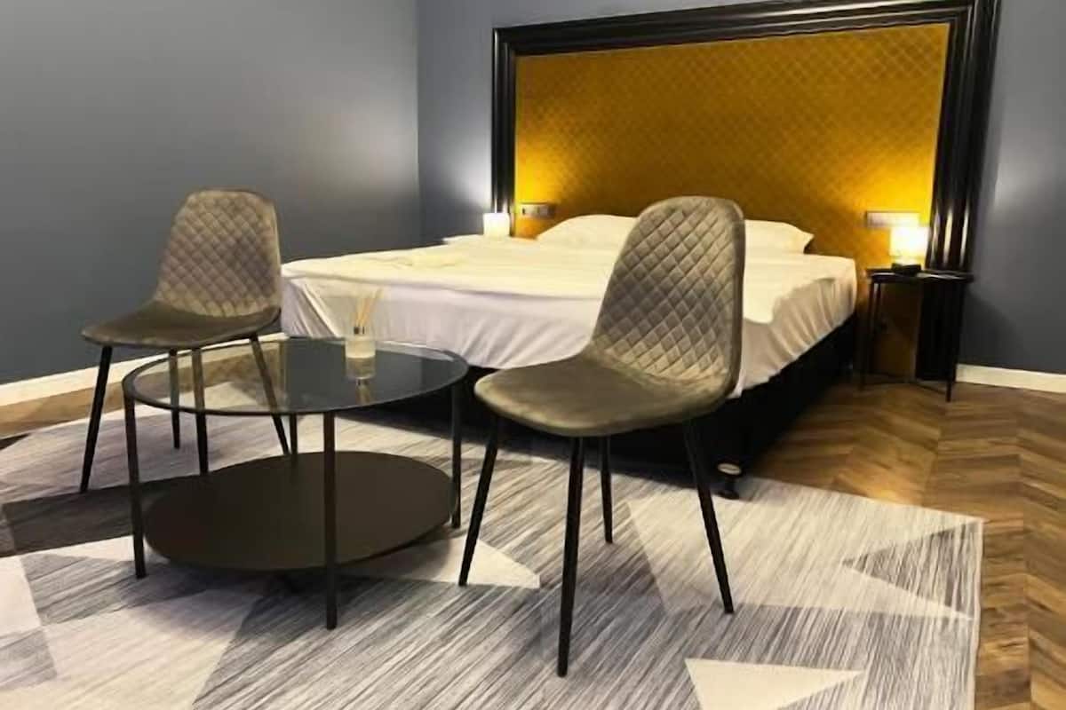 A hotel room with a bed, two chairs, a small table, and a nightstand with a lamp.